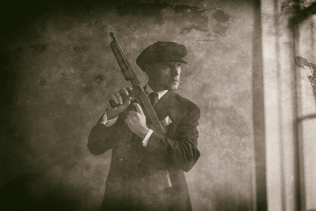 Retro 1920s english gangster wearing flat cap and suit. Standing with gun looking out window.の写真素材