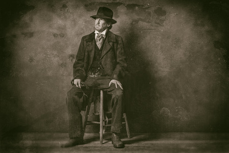 Classic wet plate photo of vintage 1900 western mature man with revolver sitting on wooden stool.の写真素材