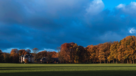 Country manor house surrounded by orange autumn trees under dark stormy sky.の写真素材