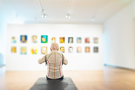 Senior woman sits on bench in white museum room with collection of artworks on wall. Rear view.の写真素材