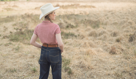 Vintage 1950s cowgirl in jeans and checkered shirt in field. Rear view.の写真素材