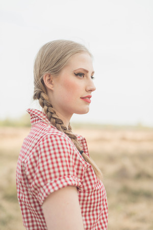 Retro 1950s young woman with braids in red checkered shirt and in countryside. Side view.の写真素材
