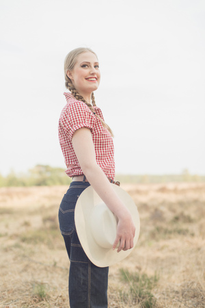Smiling retro 1950s cowgirl with white hat in red checkered shirt and blue jeans in countryside. Side view.の写真素材