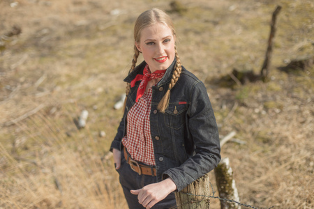 Smiling retro 1950s blonde woman in jeans leaning against wooden post in field.の写真素材