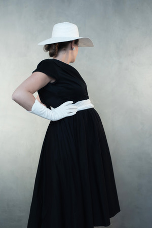 Retro 1950s pregnant woman in white hat and black dress. Looking away from camera.の写真素材