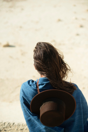 Brunette woman with brown hat in blue cloth standing in windy desert. Rear view.の写真素材
