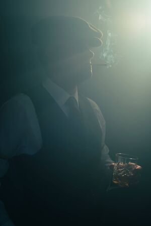 Retro businessman with cap smokes cigarette in smoky room.の写真素材