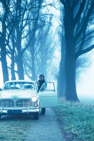 Man with pistol in black jacket standing behind door of classic car during misty dusk.の写真素材