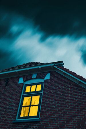 Intruder with handgun in eerie house with illuminated window under stormy sky at dusk.の写真素材