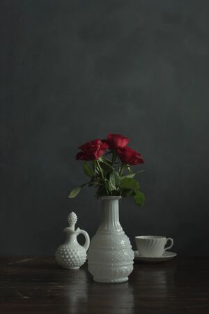 Antique white vase with red roses, tea cup and milk jug on wooden table.の写真素材