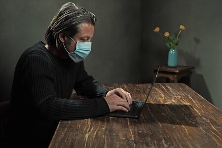 Man with hygienic mask behind laptop working at home. Side view.の写真素材