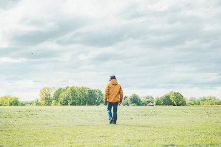 Man in yellow jacket and jeans walks in meadow in countryside.