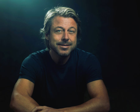 Smiling blonde man wearing black t-shirt in front of dark background looking at camera.