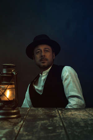 Retro cowboy victorian style man behind wooden table with historical oil lantern.の写真素材