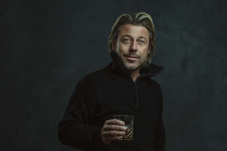Happy blond man in black woolen sweater holding glass of whiskey.の写真素材