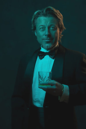 Man in tuxedo and bow tie holding a glass of whiskey. Lit by blue and orange light in front of a dark wall.の写真素材