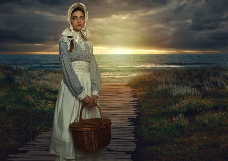 Victorian girl with hood and wicker basket in white dress and blue striped blouse on the coast at...