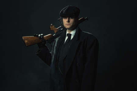 Threatening vintage man in a dark suit and cap in front of a dark wall holds a shotgun over his shoulder.の写真素材