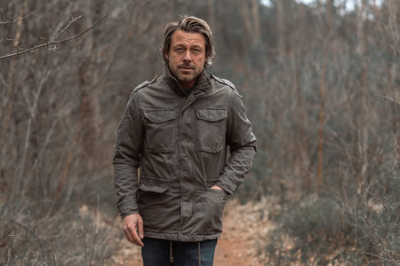 Blond man in green coat walks on a pathway between trees in nature reserve.