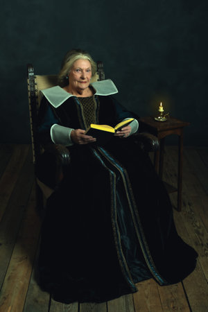 Senior woman dressed in Renaissance attire with a book sitting in a chair by a small table topped by...
