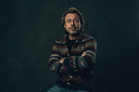 Casual blond man with stubble beard and arms crossed in cardigan sweater and jeans in front of a...