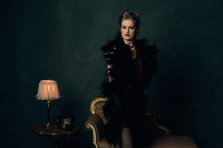 Seductive young woman with a black boa in retro boudoir attire with necklaces standing by an antique...