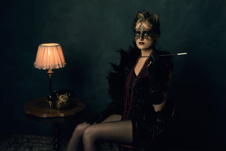 Mysterious young woman with a black boa and mask in retro boudoir attire with necklaces sitting with...