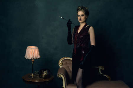 Seductive young woman with a cigarette in retro boudoir attire with necklaces stands by an antique sofa next to a coffee table with a table lamp and a telephone.の写真素材