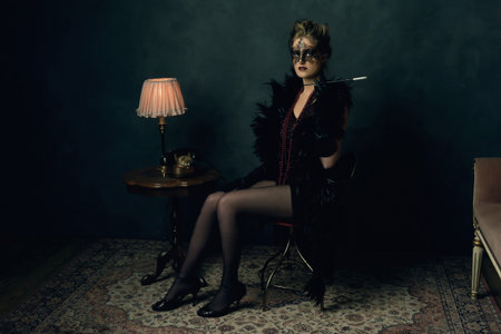 Mysterious young woman with a black boa and mask in retro boudoir attire with necklaces sitting with...