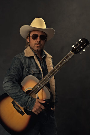 Man in denim jacket, white cowboy hat and aviator sunglasses with acoustic western guitar in front...
