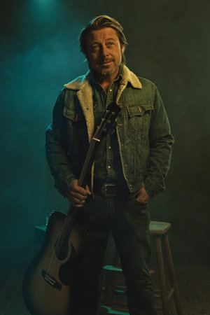 Country and western musician in jeans and a denim jacket stands with an acoustic guitar in front of...