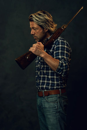 Man with blond hair in lumberjack shirt and jeans is holding a rifle over his shoulder.