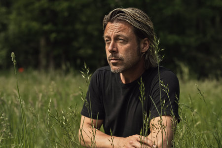 Blond man with stubble beard in a black t-shirt in a meadow with tall grass.