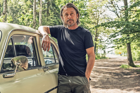 Man in a black t-shirt standing by a classic car on a forest dirt road.