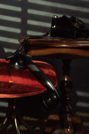 Vintage black telephone on a wooden table with the receiver on a red chair in a room with evening light shining through blinds.の写真素材