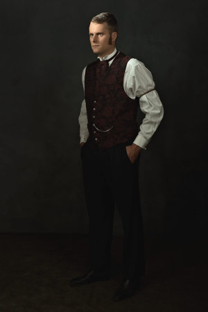 Shadowy portrait of a young man in vintage Victorian attire in a room in front of a dark gray wall.の写真素材