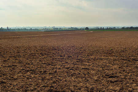 Bare arable field with a skyline of trees and a city under a cloudy sky.の写真素材