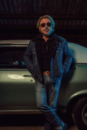 Man with blond hair and sunglasses in a jeans jacket stands by an American classic muscle car.の写真素材