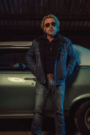 Man with blond hair and sunglasses in a jeans jacket stands by an American classic muscle car.の写真素材