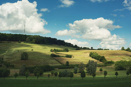 Farmland with trees and hedges in a rolling landscape under a blue cloudy sky.の写真素材