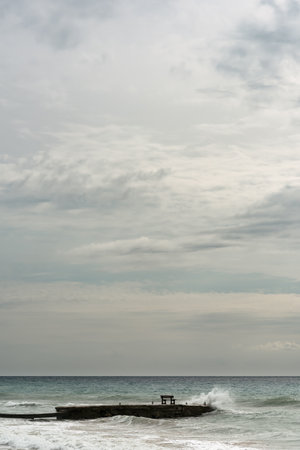 Pier with bench in splashes of wave in ocean under a cloudy sky.の写真素材