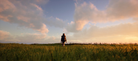 Little boy with backpack walks in pasture in countryside under a blue cloudy sky during sunset. 3D...