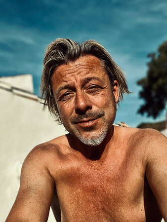 Satisfied man with blond hair and gray stubble beard enjoying the sun in backyard by white house.の写真素材