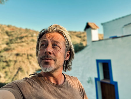 Happy blond man with stubble in brown t-shirt by a white house in sunny countryside.の写真素材