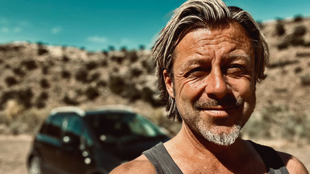 Tanned blond man with beard in singlet shirt in arid sunny environment with black car in background.