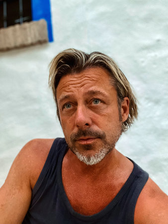 Tanned man with blond hair, blue eyes and gray stubble beard in blue singlet shirt outdoor by a...