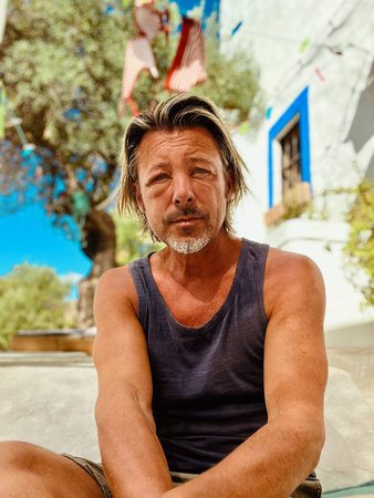 Tanned blond man with gray stubble beard in singlet sitting outside by a spanish white house.