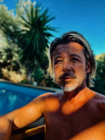 Tanned rugged blond man with gray stubble beard shirtless outside by a swimming pool.