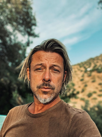 Tanned man with blond half long hair and gray stubble beard in brown t-shirt enjoys sunny summer...
