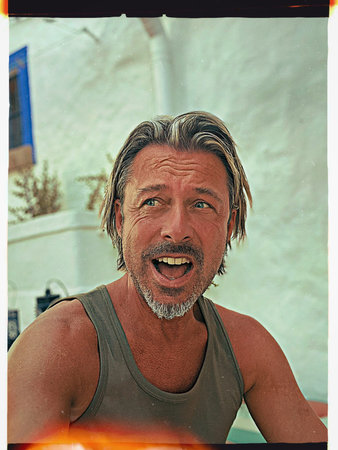 Screaming tanned blond man with beard in green singlet shirt sits by white stucco wall outside.